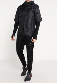 Homme portant une veste de sport noire Nike à manches courtes sur des manches longues, un pantalon Nike noir et des chaussures de course noires, debout devant un fond blanc.