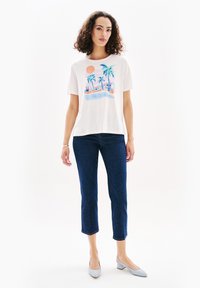 Camiseta blanca de algodón con un gráfico colorido de palmeras y el sol. Combinada con unos jeans cortos de color azul oscuro y zapatos de tacón azul claro.