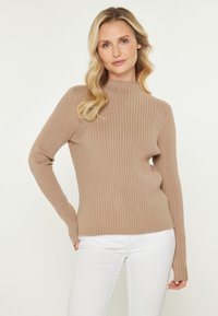 Beige gerippter Strickpullover mit hohem Kragen und langen Ärmeln, der eine taillierte Passform und eine weiche Textur aufweist. Kombiniert mit weißen Hosen.