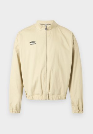 Beige glidelåsjakke med ståkrage, elastiske mansjetter og nederkant, med en liten svart Umbro-logo på venstre bryst.