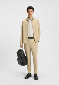 Beige gestructureerde jas over een licht overhemd, gecombineerd met beige broeken. Een zwarte rugzak en schoenen maken de look compleet, met eenvoudige hardware.