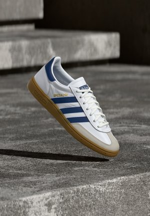 Бял спортен обувка Adidas Spezial с сини ивици и бежова подметка, плаваща над сиви бетонни стъпала.