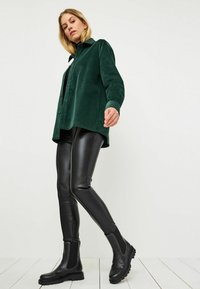 Green corduroy shirt, relaxed fit, button front. Black faux leather leggings, smooth texture. Black ankle boots with elastic side panels and studs.