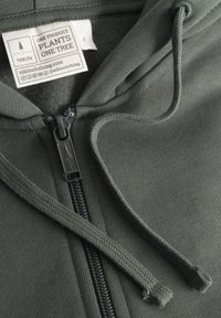 Sudadera verde oscuro con cremallera frontal, tejido texturizado y cordones ajustables. Incluye una etiqueta que indica producción sostenible.