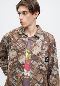 Barna álcázó gombos ing levélmintákkal, elülső zsebbel és fekete logóval, egy szürke grafikás póló felett viselve.