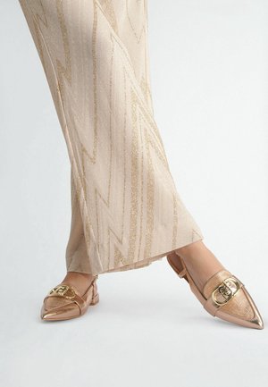 Zapatos slingback en tono oro rosa, con punta afilada y textura metálica, adornados con hebillas doradas, combinados con pantalones de tela beige con estampado.