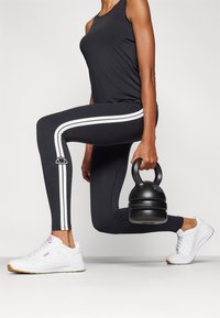 Melni treniņbikses ar baltām sānu sloksnēm, kombinētas ar melnu kettlebellu un baltiem sporta apaviem. Augšdaļa ir melna bez piedurknēm.