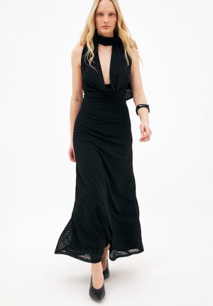 Donna che cammina con un vestito lungo nero senza maniche con pois trasparenti, scollatura profonda, collo a sciarpa, tacchi neri e braccialetto nero.