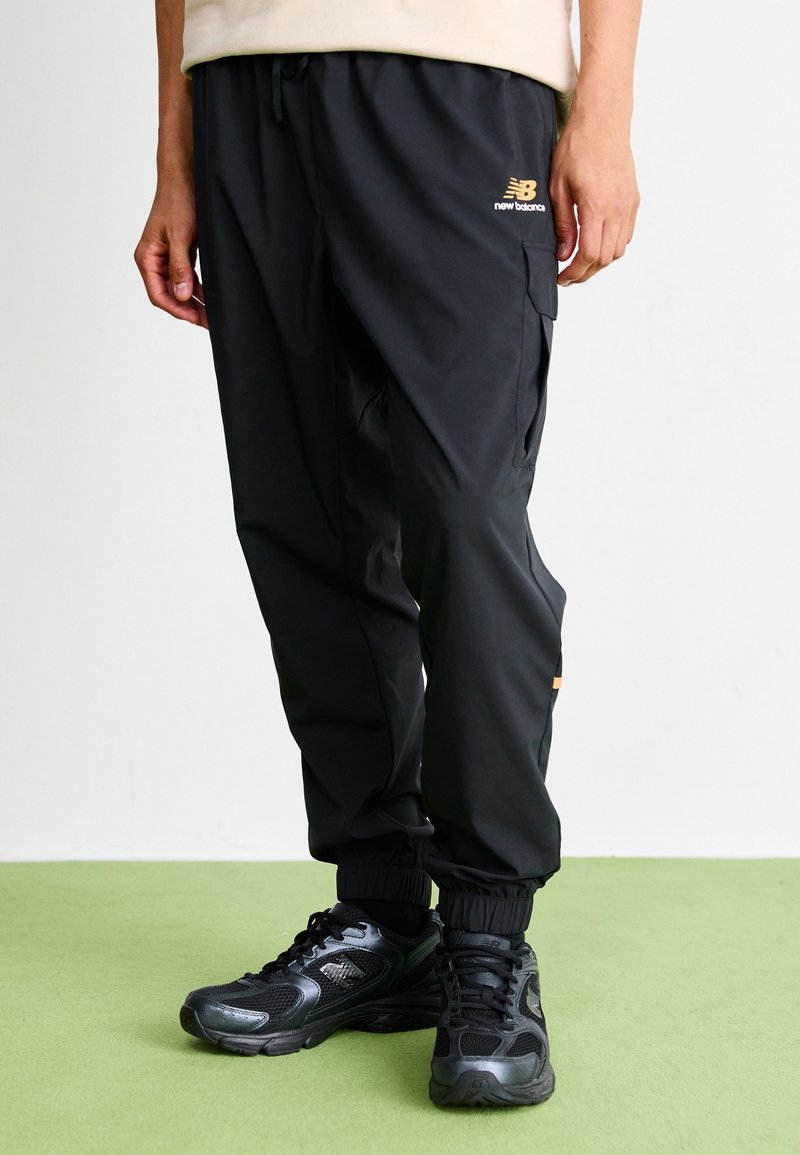 Person wearing black New Balance cargo pants and black athletic shoes, standing on green floor against a white wall.