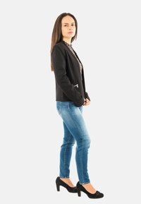 Femme se tenant de profil, portant un blazer noir, un blouse à motifs, un jean bleu et des talons noirs sur un fond clair et uni.