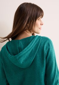 Teal hoodie van zachte stof, met een trekkoordkap, ribgebreide boorden en een ontspannen pasvorm. De textuur ziet er warm en gezellig uit.