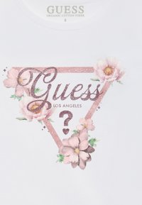 Glitrende rosa trekantet Guess-logo med teksten "Los Angeles", omkranset av rosa og hvite blomsterdesign på hvitt stoff.