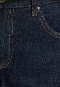 Tela de mezclilla azul oscuro de los jeans con costuras doradas. Presenta un bolsillo frontal, cierre con botón y un acabado texturizado.