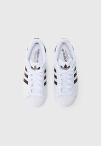 Białe sportowe trampki z trzema brązowymi paskami, teksturowanym noskiem i białymi sznurowadłami. Logo Adidas na języku i boku.