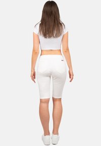 Top court blanc, shorts blancs jusqu'aux genoux, vue arrière. Les shorts présentent un design à cinq poches, avec des ourlets retroussés. Baskets blanches visibles.