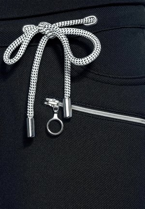 Gros plan sur un pantalon noir avec un cordon de serrage blanc et noir noué en nœud au-dessus d'une tirette de fermeture éclair argentée en forme d'anneau et d'une fermeture éclair blanche.