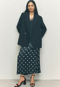 Sacou negru cu dublă deschidere peste un top verde, asortat cu o fustă bleumarin cu un model cu puncte albe și sandale cu toc bloc negre.