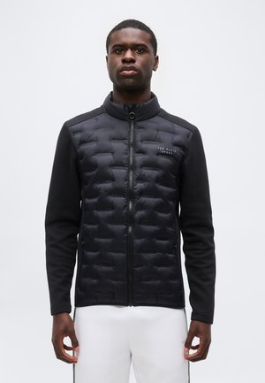 Uomo che indossa una giacca trapuntata nera con zip e logo "Ted Baker Sport", in piedi davanti a uno sfondo chiaro e uniforme.