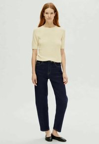 Puha bézs, rövid ujjú pulóver sötétkék, magas derekú farmernadrággal párosítva. Mindkét ruhadarab laza szabású; a szettet fekete lapos cipők egészítik ki.