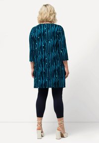 Tunique bleue à motifs avec des lignes verticales, présentant une coupe ample et des manches trois quarts, associée à des leggings navy et des talons blocs clairs.