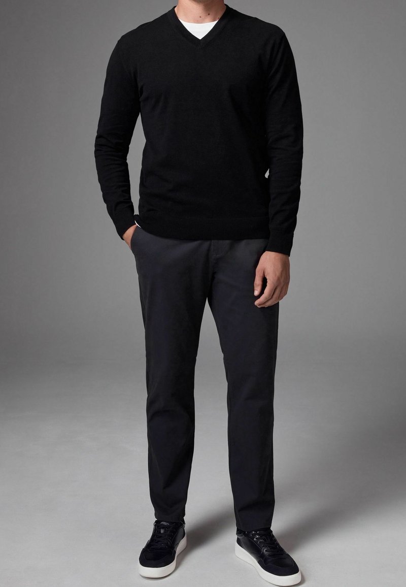 Homme portant un pull noir à col en V, un pantalon foncé et des baskets noires à semelles blanches, posant avec une main dans la poche sur fond gris.