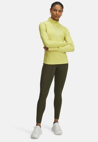 Yellow long-sleeve athletic top with a half-zip collar, paired with dark green leggings and white sneakers. Features smooth, fitted design.
