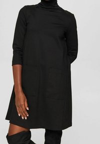 Robe noire à manches longues avec col roulé et poches avant, portée avec des bottes noires montantes assorties, vue des épaules jusqu'à mi-cuisse.