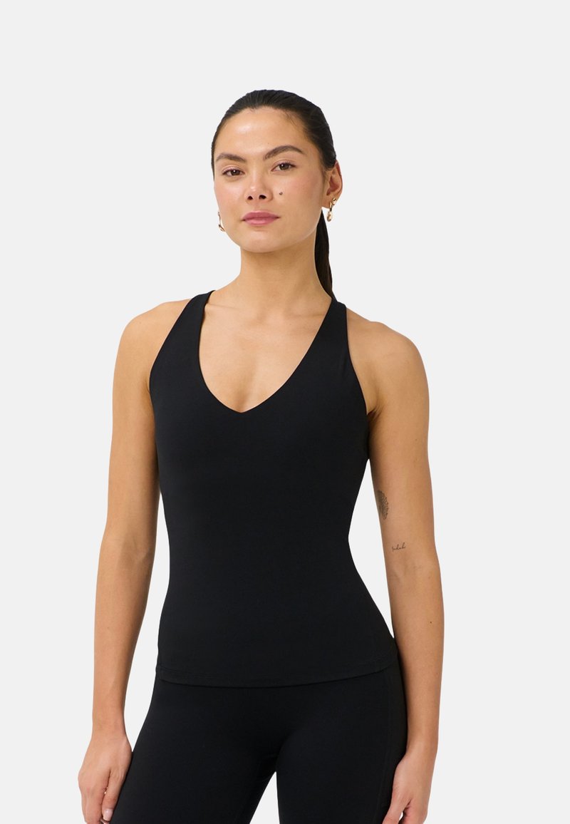Woman with dark hair tied back wearing a black sleeveless V-neck top and black leggings, standing against a plain light background.