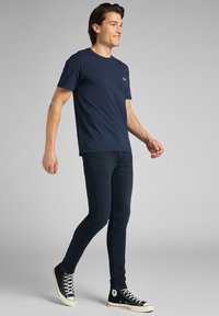 Maglietta a maniche corte blu navy in tessuto morbido, indossata con jeans scuri slim-fit e sneakers alte nere con suola bianca.