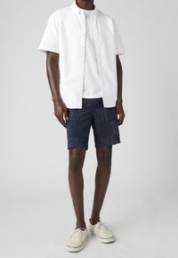 Hombre vistiendo una camisa blanca de botones de manga corta sobre una camiseta blanca, pantalones cortos de rayas azules y beige, y zapatillas beige, de pie sobre un fondo gris claro.