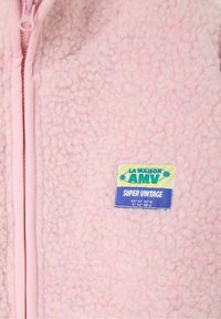 Chaqueta de forro polar rosa difusa con cremallera frontal y una colorida etiqueta "LA MAISON AMV SUPER VINTAGE" cosida en el pecho.