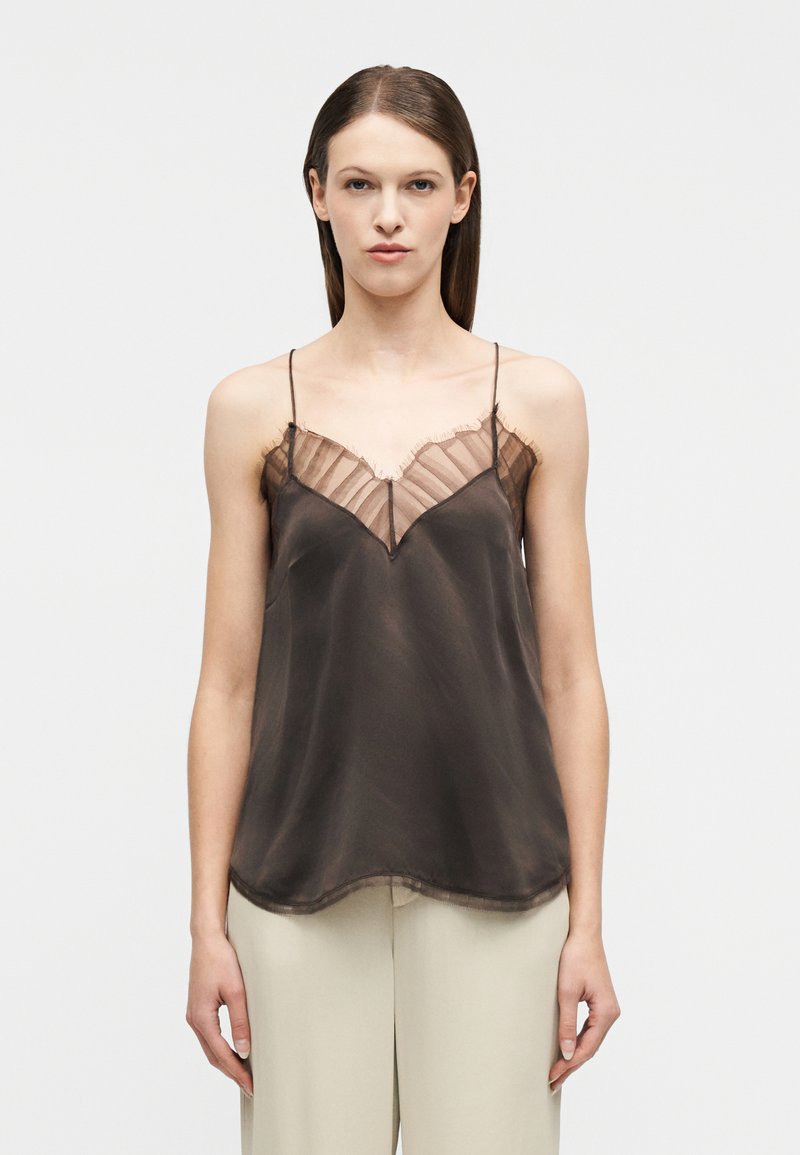 Γυναίκα που φοράει διαφανές καφέ σ κούρο τοπ με βολάν και λεπτές τιράντες, συνδυασμένο με ανοιχτό μπεζ παντελόνι, στέκεται μπροστά σε λευκό φόντο.