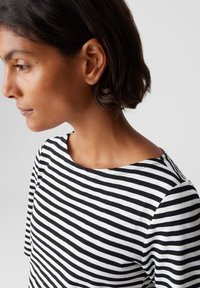 Femme aux cheveux courts et foncés portant une chemise rayée noire et blanche, tournée vers la gauche devant un fond clair.