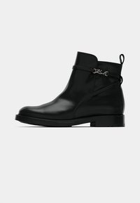 Bottines en cuir noir avec un petit talon, détail de sangle latérale et logo « Karl » en argent sur une surface lisse et polie, le tout sur fond blanc.