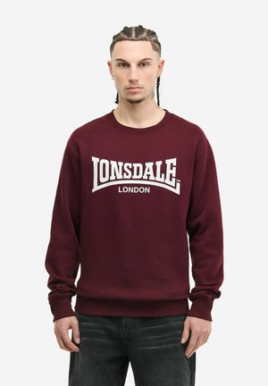 Mladić s pletenicama, odjeven u bordo Lonsdale London majicu i tamne traperice, stoji ispred jednobojne bijele pozadine.