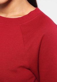 Rode sweatshirt met een geribbelde nek en textuurstof, die een gladde afwerking laat zien met subtiele stiksels langs de schoudernaad.