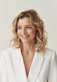 Balts lina bleizers ar dziļo V-izgriezumu, strukturētām plecu daļām un lieliem pogām; modelim ir viļņaini, vidēja garuma mati un viegls smaids.