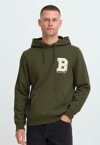 Hombre con cabello corto que lleva una sudadera verde oliva con una gran letra blanca "B" en el pecho y pantalones negros, de pie con una mano en el bolsillo.