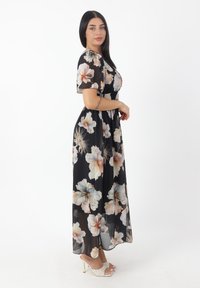 Robe maxi avec un fond noir et un grand imprimé floral dans des couleurs pastel. Elle a des manches courtes et une taille cintrée. Portée avec des talons légers.