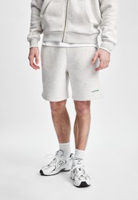 Lys grå bomullshoodie med glidelås foran, kombinert med matchende grå shorts. Utsmykket med grønn merking på shortsen og hvite treningssko.