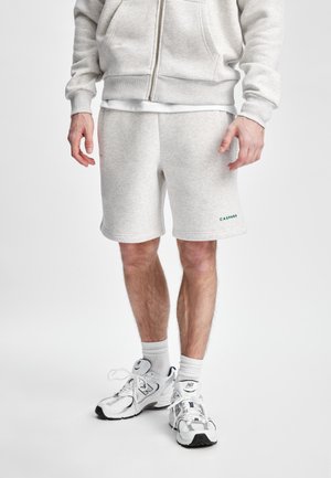 Lys grå bomullshoodie med glidelås foran, kombinert med matchende grå shorts. Utsmykket med grønn merking på shortsen og hvite treningssko.
