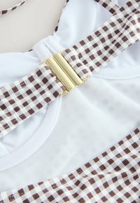 Bretelle de bikini en tissu à carreaux marron et blanc avec une boucle en métal doré sur fond en maille blanche et tissu uni.