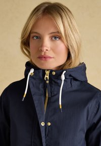 Blonde Frau mit blauen Augen, die eine marineblaue Kapuzenjacke mit weißen Kordeln und goldenen Reißverschlussdetails trägt, blickt nach vorne vor neutralem Hintergrund.