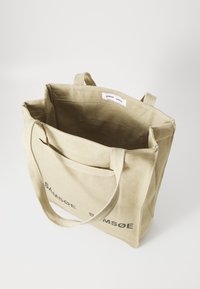 Canvas tote bag in light beige with dual handles, featuring a simple design and logo printed in black on the front. Interior view includes pockets.