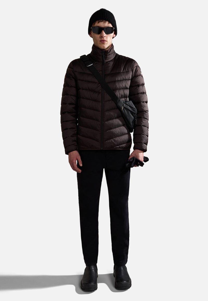 Mann trägt eine schwarze Mütze, Sonnenbrille, dunkelbraune Steppjacke, schwarze Hose, schwarze Stiefel, hält schwarze Handschuhe und hat eine schwarze Umhängetasche.