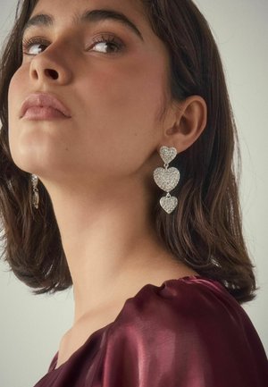 Woman with short dark hair wearing three-tiered sparkling heart-shaped earrings and a burgundy satin top, looking slightly upward.