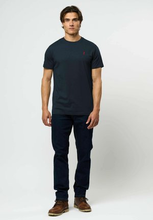 Marineblaues Baumwoll-T-Shirt mit kurzen Ärmeln und rundem Ausschnitt, mit einem kleinen roten Emblem auf der linken Brust, kombiniert mit dunklen Jeans und braunen Stiefeln.