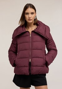 Giacca a vento borgogna con grande colletto, design trapuntato, chiusura con zip e tasche laterali. Indossata sopra un completo nero.