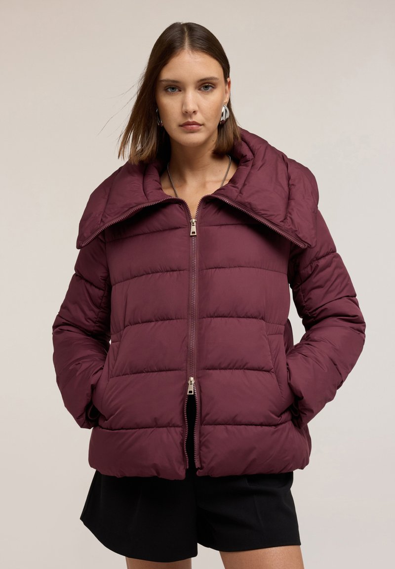 Giacca a vento borgogna con grande colletto, design trapuntato, chiusura con zip e tasche laterali. Indossata sopra un completo nero.