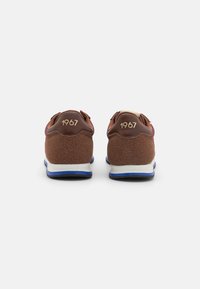 Zapatillas deportivas marrones con parte superior de ante texturizado, suela azul y detalle dorado "1967" en la parte trasera. Diseño minimalista con acentos en contraste.